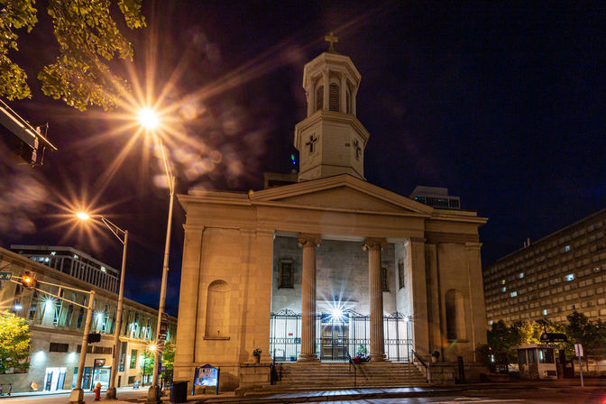 Neoclassical church with tall columns and an illuminated steeple topped by a cross on a downtown city street at night, glowing streetlights creating starburst light effects.