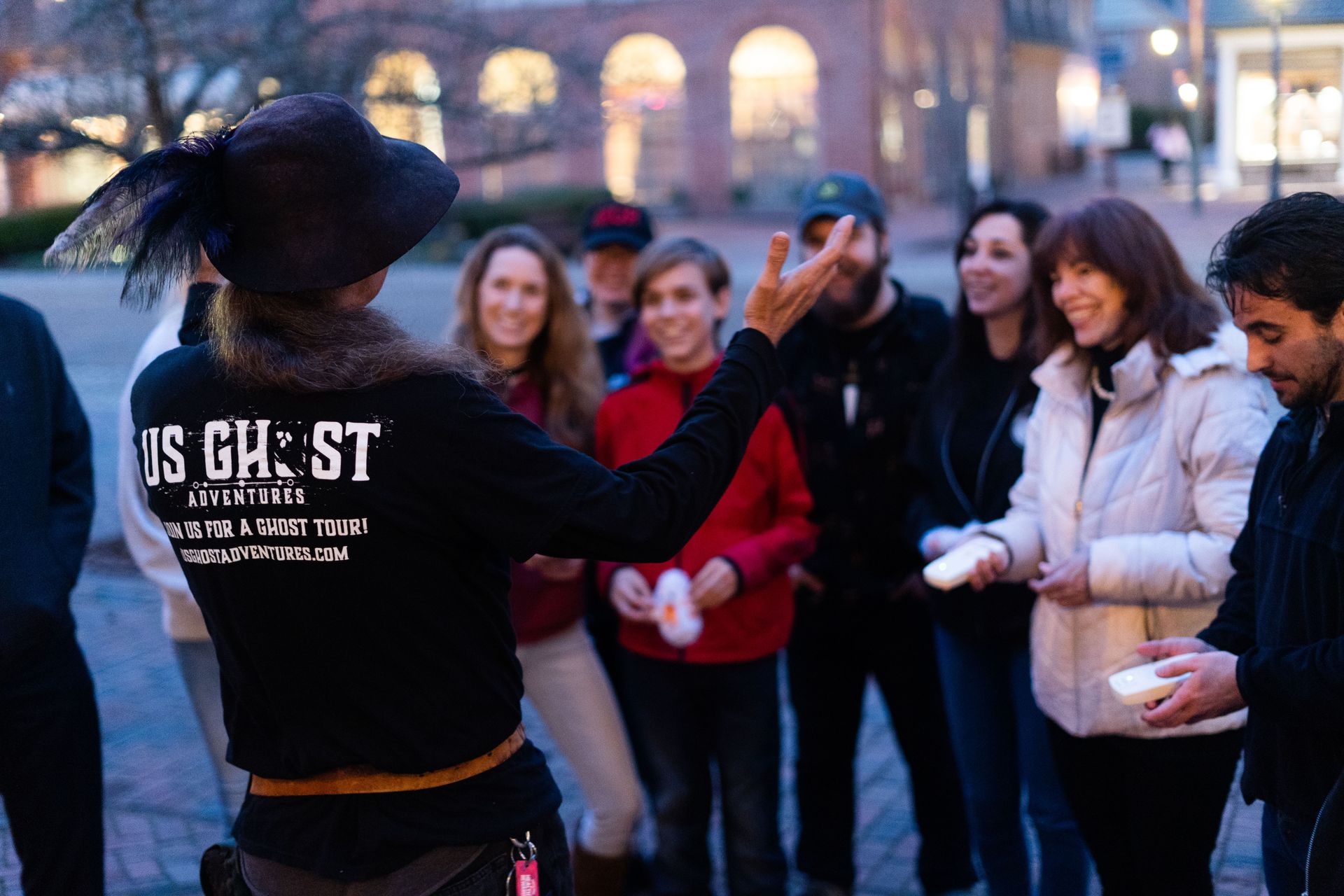 Tour guide wearing a feathered hat leading a lively ghost tour at dusk in a brick downtown square, a smiling group holding small handheld devices.