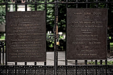 Ornate wrought-iron gate with two weathered bronze plaques and a small padlock marking a historic 17th-century burial ground in Boston, leafy cemetery visible beyond the bars.
