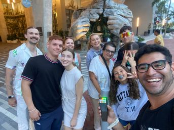 Group selfie of smiling people in an urban plaza outside a hotel entrance with a giant stone hands sculpture, palm trees and lit sidewalk — fun vacation photo-op vibe.