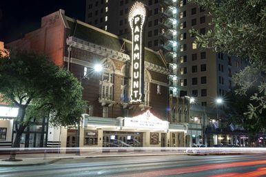 Vintage brick theater with glowing vertical marquee on a bustling downtown street at night, car light trails and a nearby high-rise building