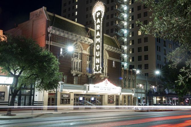 Vintage brick theater with glowing vertical marquee on a bustling downtown street at night, car light trails and a nearby high-rise building