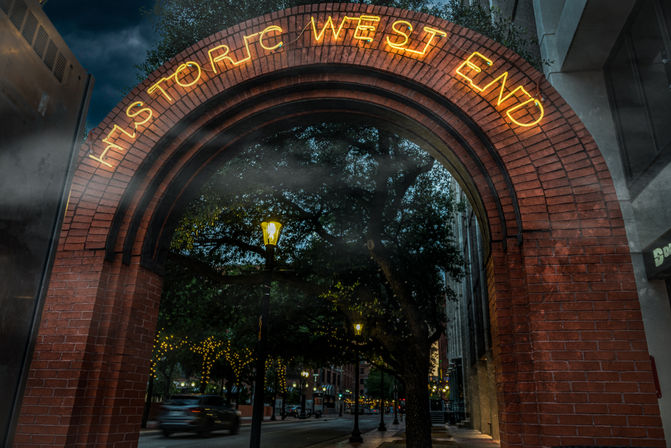 Neon “Historic West End” brick arch framing a tree-lined, lamp-lit street at dusk with a moving car blur and twinkling string lights