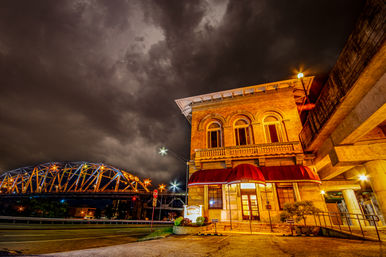 Historic brick building with red awning lit at night on a riverfront street, glowing steel bridge in the background under dramatic stormy clouds — downtown urban nightscape.