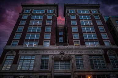 Low-angle view of a towering historic red-brick courthouse in downtown Dallas at dusk, glowing windows beneath dramatic purple-pink clouds.
