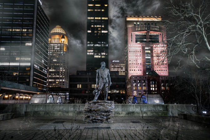 Moody nighttime downtown plaza with a bronze statue on a stacked-rock pedestal framed by illuminated skyscrapers and stormy clouds.