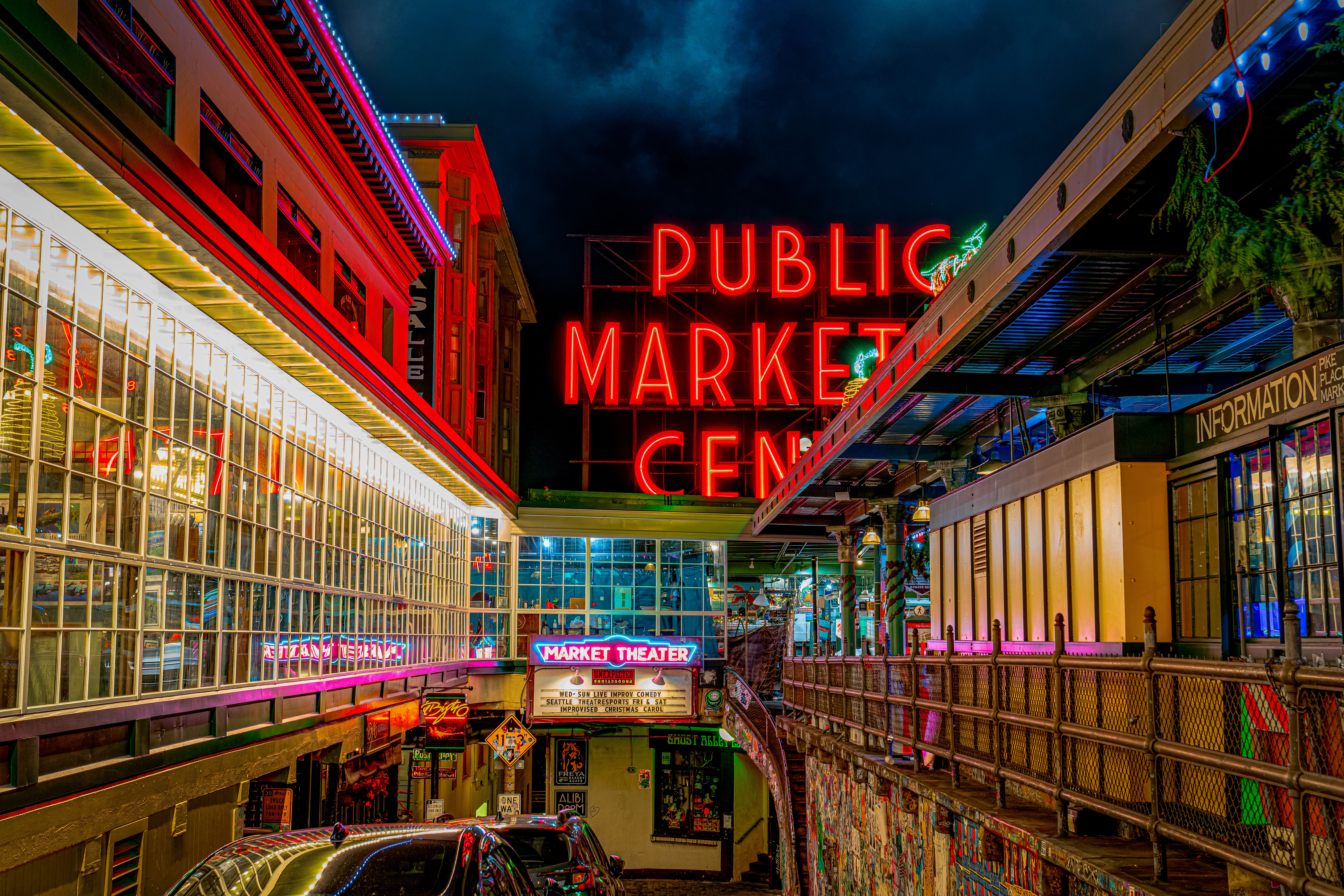 Neon-lit Seattle market at night — large red "PUBLIC MARKET" sign above glowing glass storefronts, a vintage theater marquee, colorful overhead lights, pedestrian walkway and parked cars.