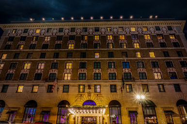 Historic downtown hotel facade at night — ornate brick with white decorative trim, rows of warm-lit windows, illuminated marquee entrance, flags and streetlamp.