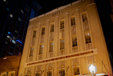 Nighttime view of a historic downtown multi-story brick storefront with tall vertical windows, ornate early-20th-century facade and vintage marquee glowing under warm streetlights in an urban scene.
