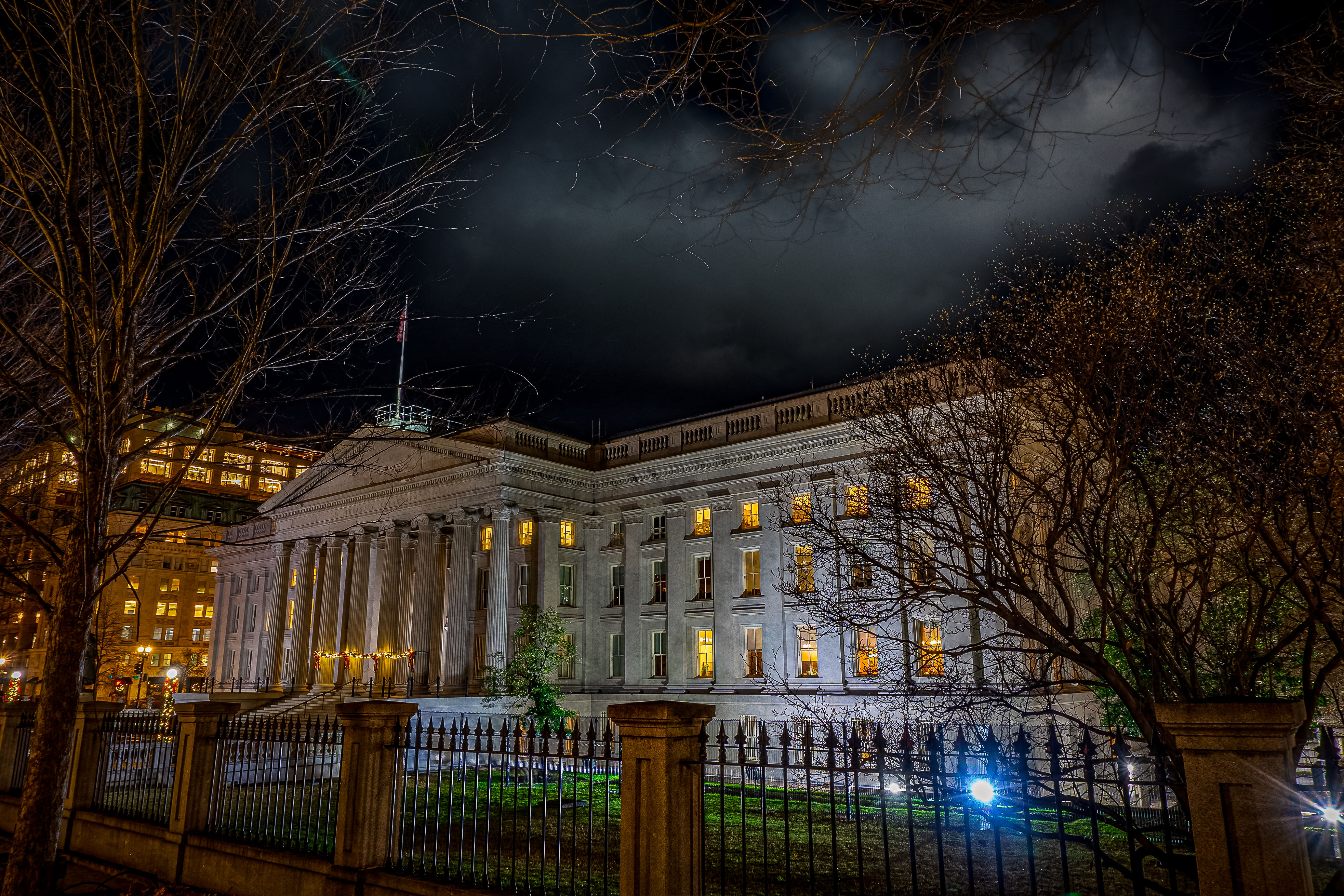 Night view of a grand neoclassical government building in a downtown city center, glowing windows and tall columns behind an iron fence, leafless trees silhouetted against a dramatic cloudy sky