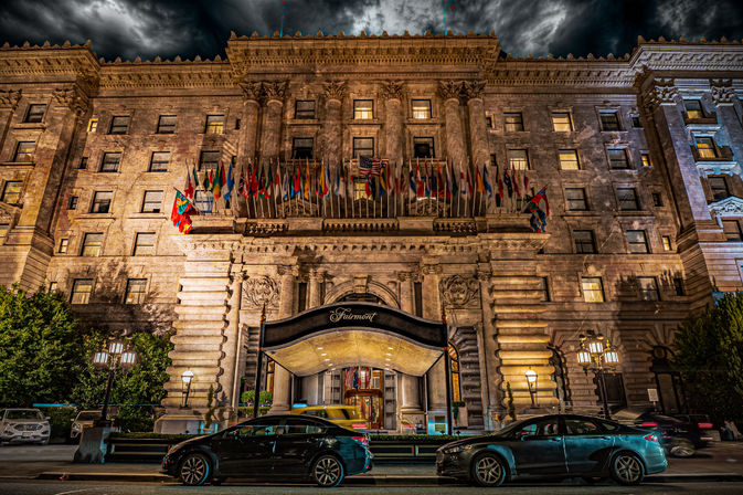 Dramatic nighttime, cinematic view of a grand historic hotel entrance with ornate stone facade and a row of international flags above a lit canopy, parked cars on the city street and stormy clouds overhead.