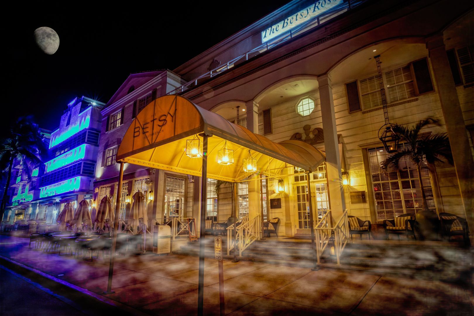 Moonlit Art Deco hotel entrance with glowing yellow canopy and hanging lanterns, neon-lit buildings and wisps of street fog along a Miami Beach sidewalk at night.
