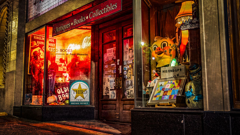 Nighttime antique bookstore storefront in a historic district with glowing red display windows showcasing vintage books, colorful toys and collectibles, an oversized cartoon pig figure, lamp and wooden entry door.