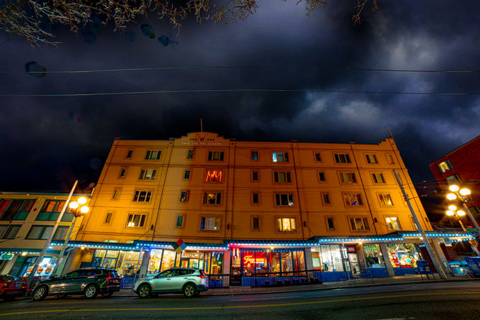 Nighttime downtown scene with a five-story yellow building, neon-lit storefronts, parked cars, glowing street lamps and dramatic storm clouds overhead.