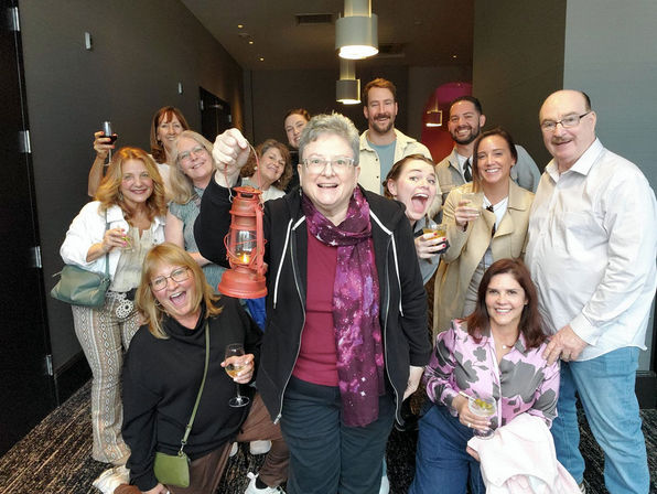 Cheerful mixed-age group posing in a hotel corridor for an indoor gathering, many holding drinks and one person holding a vintage red lantern under pendant lights.