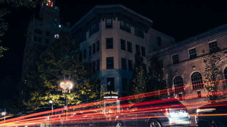 Long-exposure downtown night scene with red car light trails streaking past a historic corner building, glowing streetlamp and parked cars.