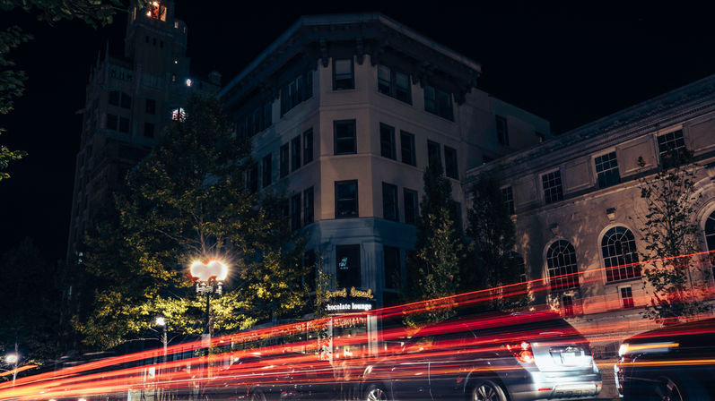 Long-exposure downtown night scene with red car light trails streaking past a historic corner building, glowing streetlamp and parked cars.