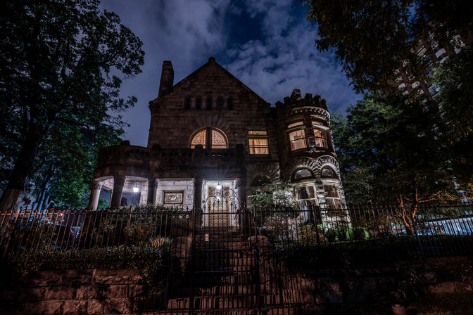 Moody nighttime view of a historic stone mansion with turret and arched windows, glowing porch lights and ornate iron gate, framed by trees under a cloudy sky.