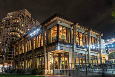 Two-story glass-and-wood urban pavilion glowing at night with warm interior lights, string lights on the exterior, balconies and modern high-rise apartments in the background