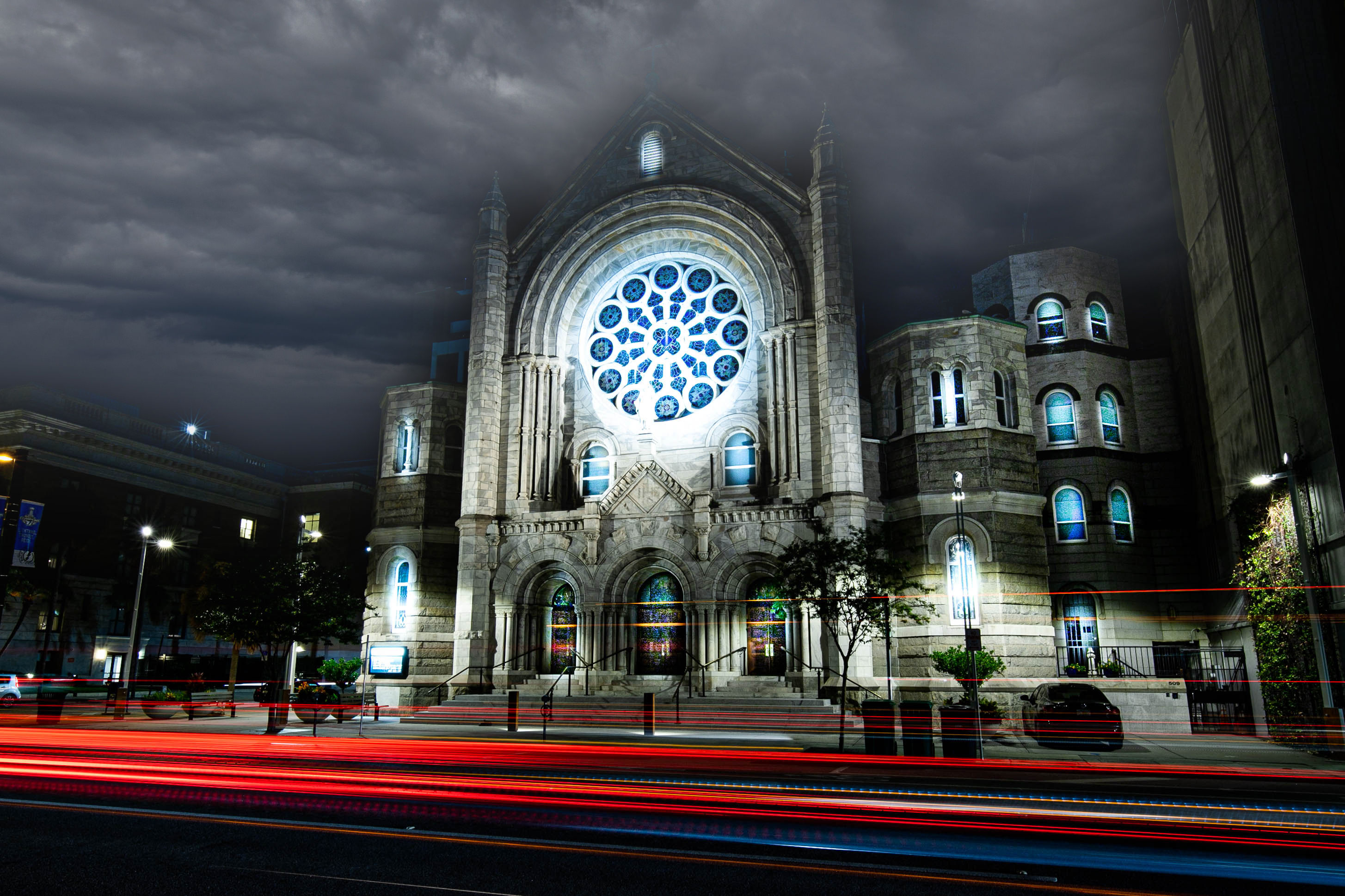 Dramatic downtown night scene of a Gothic-style stone church with a glowing blue rose window under stormy clouds, framed by trees and streaking red car light trails on the street.