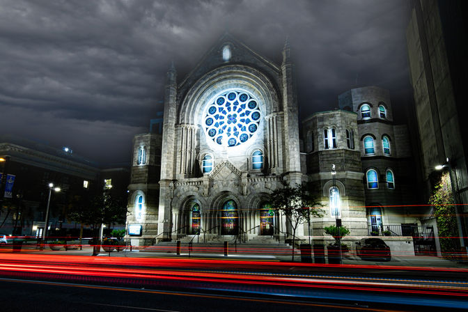 Dramatic downtown night scene of a Gothic-style stone church with a glowing blue rose window under stormy clouds, framed by trees and streaking red car light trails on the street.