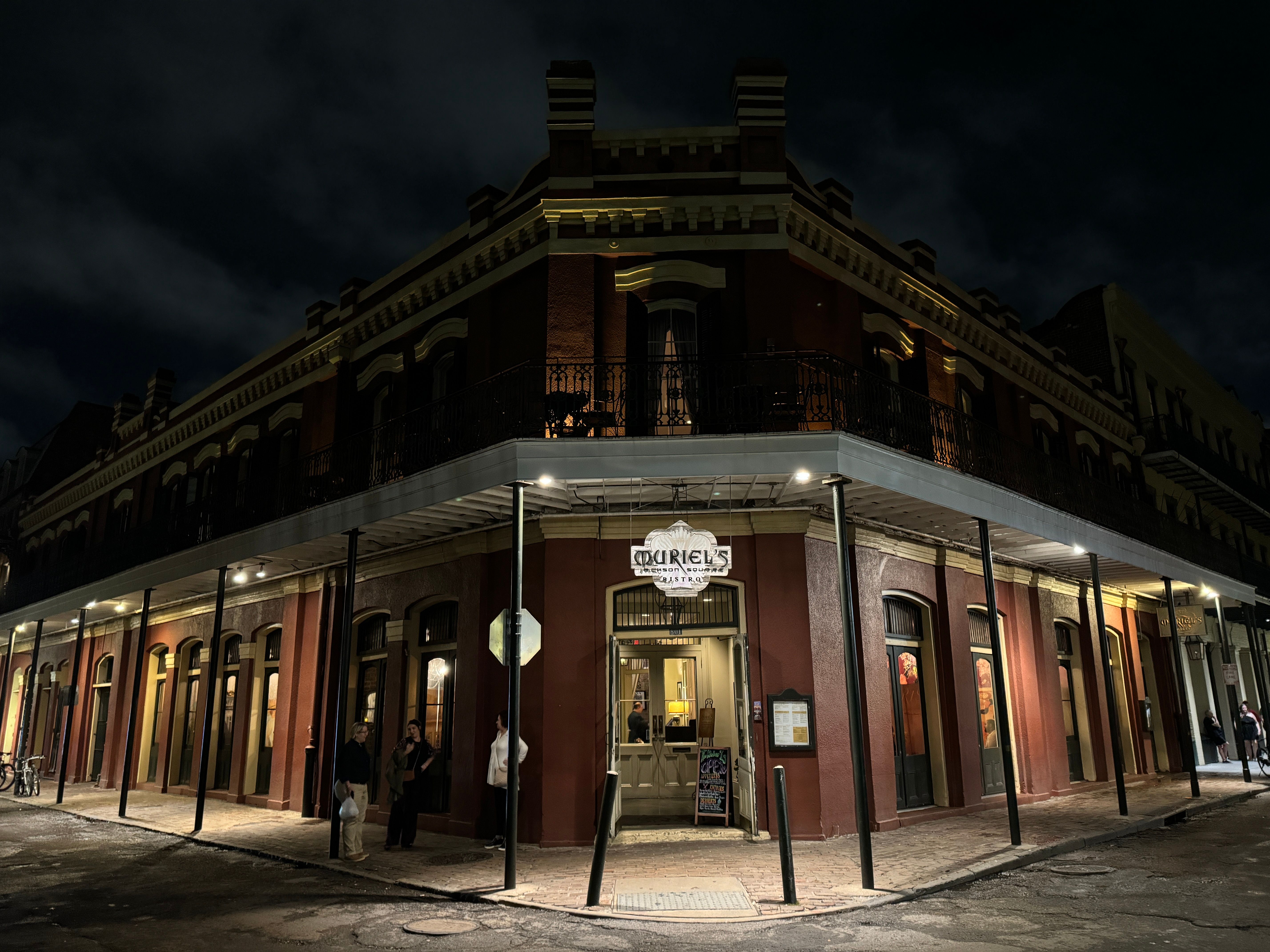 Moody night shot of a historic red brick corner building with a wrought-iron balcony, warm-lit restaurant entrance, and a few pedestrians on the sidewalk.