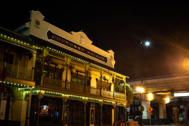 Historic two-story saloon exterior at night with ornate wooden balcony, warm string lights, and a nearby highway overpass.