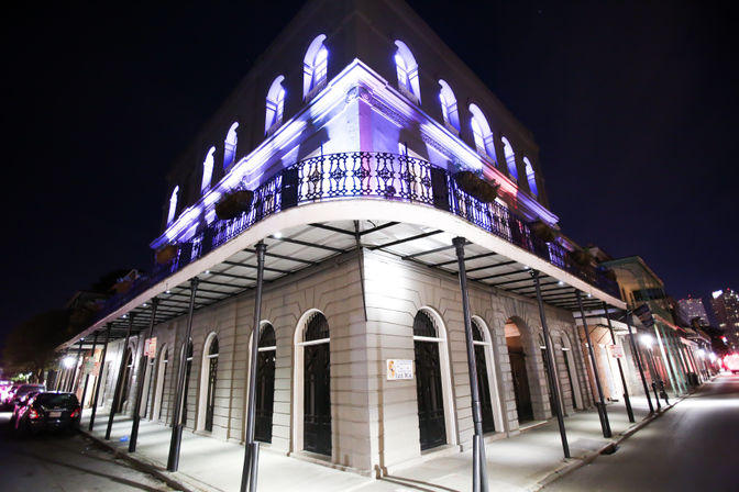 Nighttime corner of a historic French Quarter–style building in New Orleans with a glowing purple-lit wrought-iron balcony, arched ground-floor doorways and sidewalk columns.