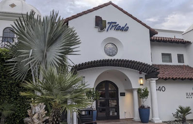 White stucco Spanish-style building with red clay tile roof, inviting arched double doors under a wooden pergola, round window above, and large fan palm plus potted plants at the entrance.