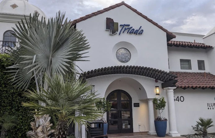 White stucco Spanish-style building with red clay tile roof, inviting arched double doors under a wooden pergola, round window above, and large fan palm plus potted plants at the entrance.
