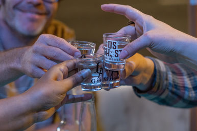 Close-up of friends clinking printed shot glasses in a cheerful cheers at a bar, clear spirits and smiling blurred faces in warm evening light.