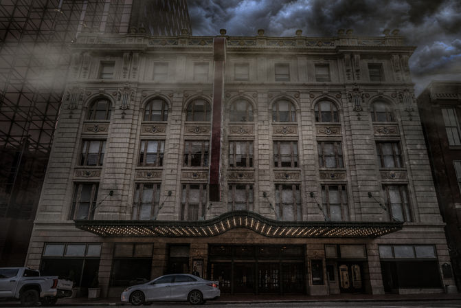 Moody downtown scene: ornate historic theater facade with arched windows and illuminated marquee lights, parked cars at the curb and stormy clouds overhead.
