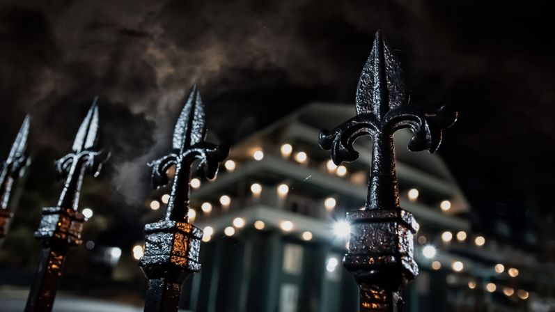Close-up of wet wrought-iron fleur-de-lis fence finials glistening at night with moody blurred building bokeh lights