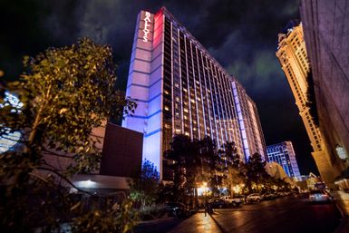Night view of a glowing high-rise hotel on the Las Vegas Strip with colorful exterior lighting, trees and parked cars along a wet street.
