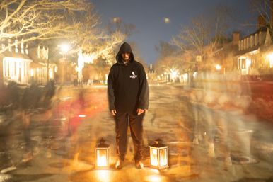 Hooded figure standing between two glowing lanterns on a lantern-lit historic street at night, long-exposure blur of passing people creating ghostly motion