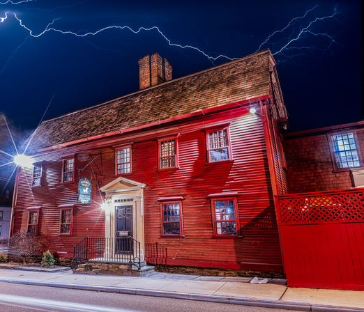 Historic red colonial-style wooden house on a quiet street at night, lit windows and porch light under dramatic lightning streaks in a deep blue sky.