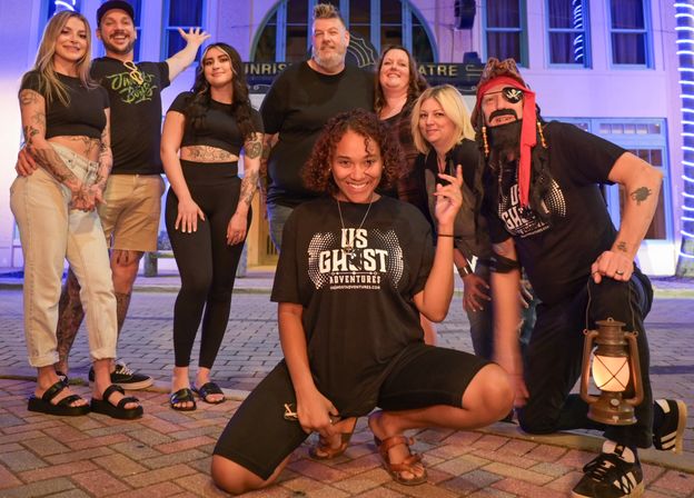 Group of eight adults posing on a neon-lit brick sidewalk in front of a small theatre at night during a ghost tour; one woman kneels front smiling while a person in a pirate costume holds a lantern.