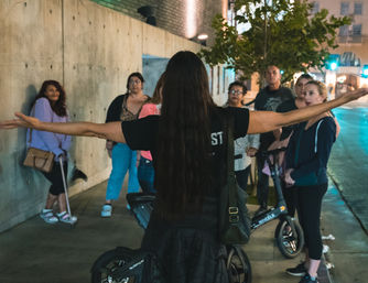 Tour guide with outstretched arms addressing a small nighttime city sidewalk group gathered around electric scooters