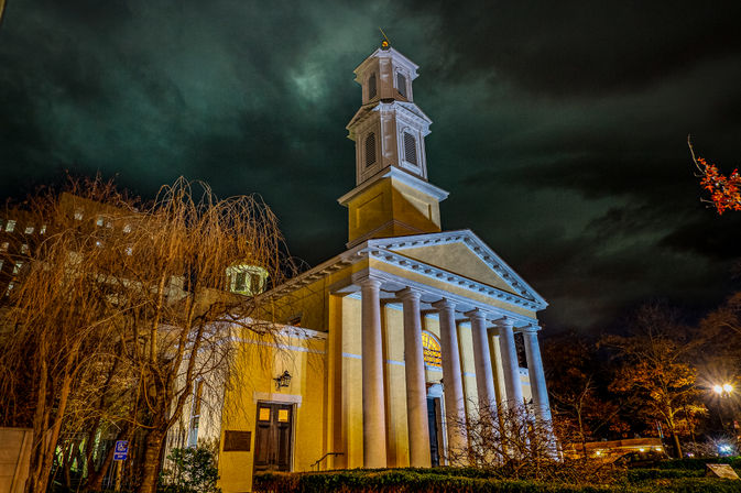 Night photo of an illuminated historic church with tall white columns and a multi-tiered steeple against a dramatic green-tinted stormy sky, framed by bare trees and city lights.