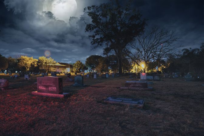 Moody suburban cemetery at night under a clouded full moon, scattered headstones and large trees lit by warm streetlights and red-tinted grass.