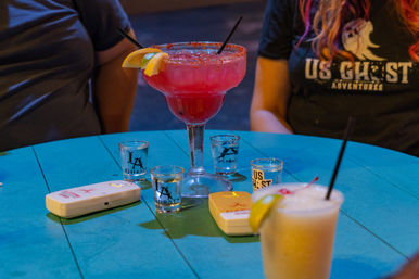 Bright pink frozen margarita in a large glass with salted rim and lemon wedge on a turquoise table, flanked by shot glasses, two order pagers and a creamy cocktail with straw, patrons seated nearby — lively bar night.