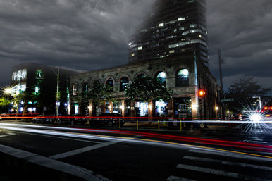 Nighttime long-exposure of a downtown brick building with arched windows and lit storefronts, streaking red and white car light trails across a crosswalk, parked cars, a bright headlight starburst, a high-rise tower and moody storm clouds overhead.