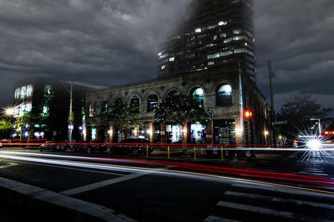 Nighttime long-exposure of a downtown brick building with arched windows and lit storefronts, streaking red and white car light trails across a crosswalk, parked cars, a bright headlight starburst, a high-rise tower and moody storm clouds overhead.