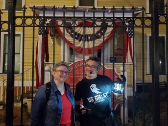 Two smiling adults at night in front of a yellow historic house draped with American bunting behind a wrought-iron fence; a ghost tour guide wearing a headset and ghost-themed shirt holds a lantern.