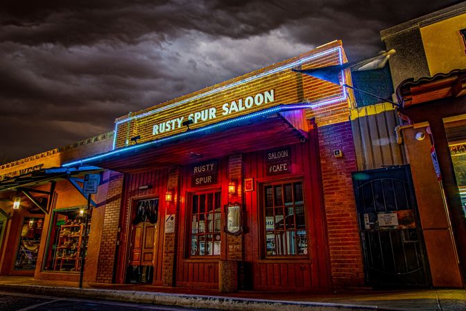 Nighttime small-town Western saloon storefront with red wood doors, brick facade, warm lanterns and blue neon trim beneath dramatic stormy clouds