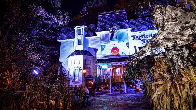 Nighttime Halloween scene of a Victorian-style haunted house bathed in purple-blue lights, pumpkin-lined steps, dried cornstalks and a tattered ghostly figure guarding the spooky entrance.