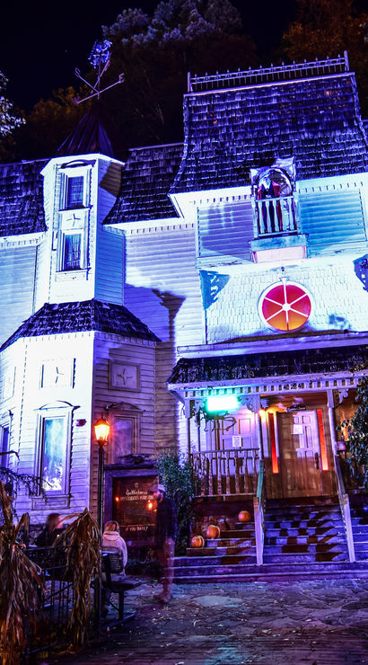 Nighttime Halloween scene of a Victorian-style haunted house bathed in purple-blue lights, pumpkin-lined steps, dried cornstalks and a tattered ghostly figure guarding the spooky entrance.