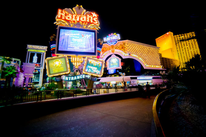 Neon-lit casino resort entrance on the Las Vegas Strip at night, glowing marquees and large digital signs above a pedestrian walkway.