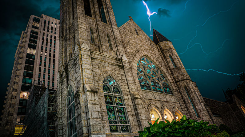 Gothic stone church with ornate stained-glass windows beside a downtown high-rise, dramatic lightning streaking across a teal night sky