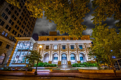 Night view of an illuminated neoclassical downtown building with grand columns and arched windows, a modern glass atrium to the left, leafy trees framing a dramatic cloudy sky.